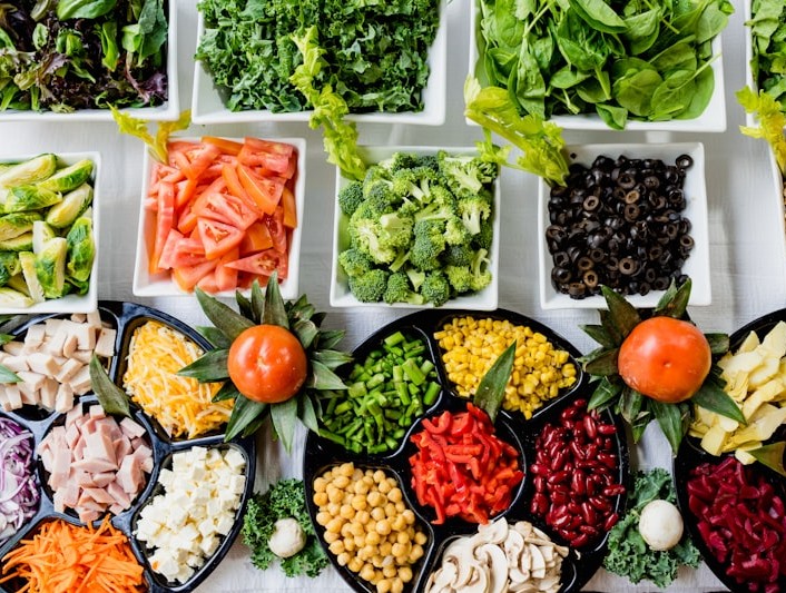 Top-down salad bar with white bowls of kale, spinach, broccoli, tomatoes, and black trays of cheese, chickpeas, and chopped vegetables